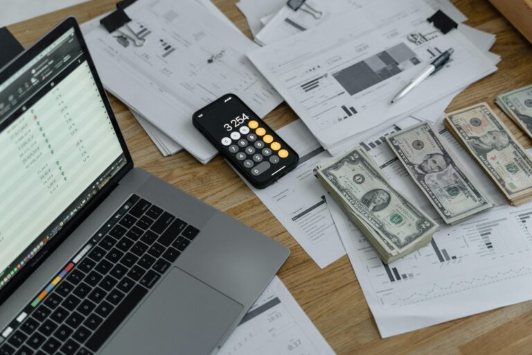A desk with financial documents, currency, dolar blue, a laptop, and phone calculator.